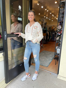 Young woman wearing a cream light-weight knit sweater with rainbow embroidery detail around the heart cut-out detailing. The sweater is styled with a light wash, distressed skinny jean and Sorel sneakers.